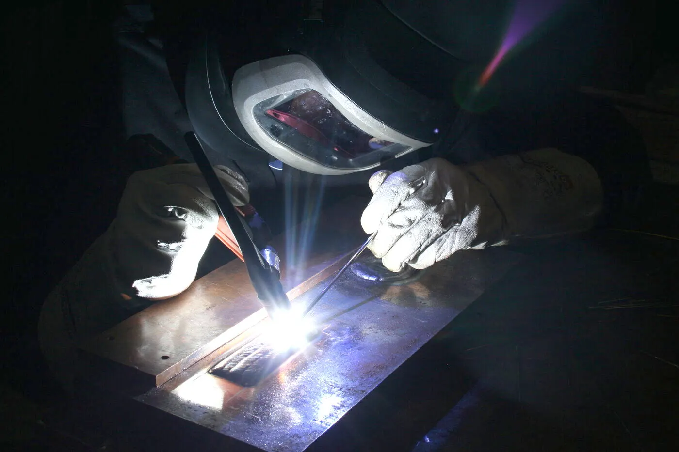 A welder wearing a protective helmet and gloves, welding a metal plate in the blue light of the arc.